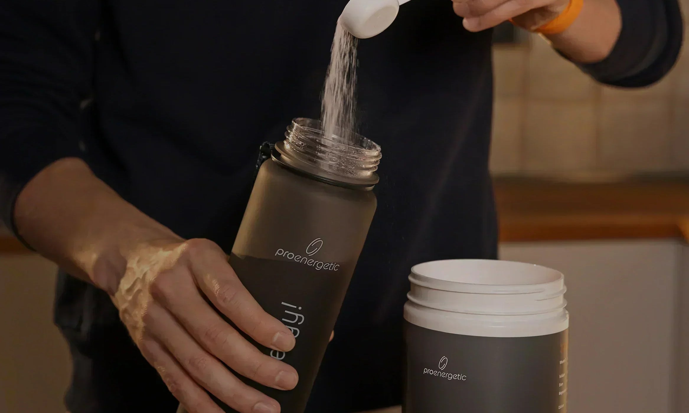 Person füllt Isomove isotonisches Pulver aus Messlöffel in schwarze Pure Move Trinkflasche neben geöffnetem Behälter