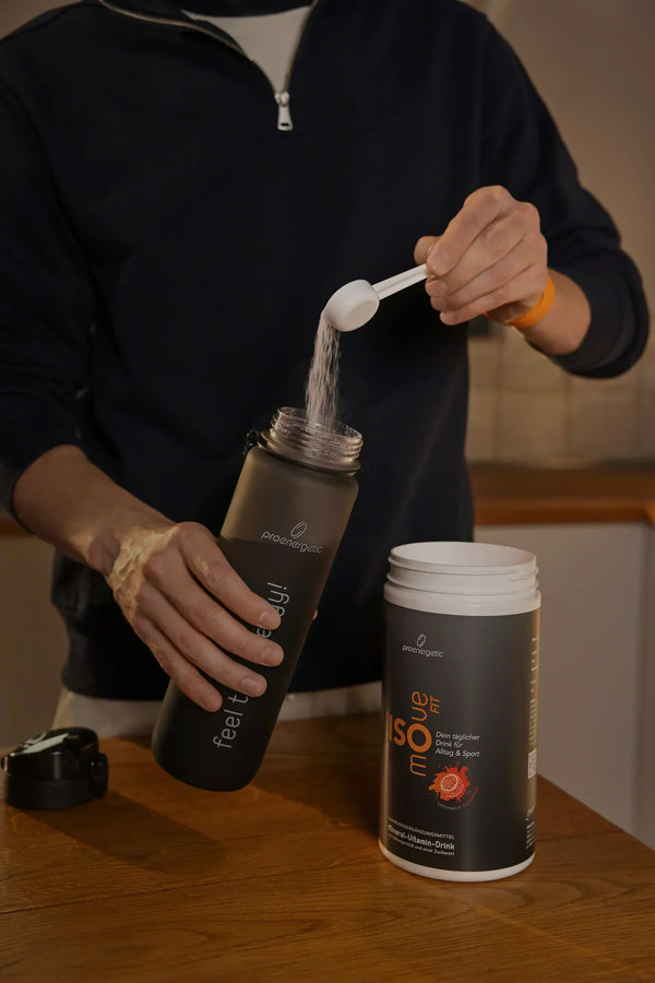 Person füllt Isomove isotonisches Pulver aus Messlöffel in schwarze Pure Move Trinkflasche neben geöffnetem Behälter