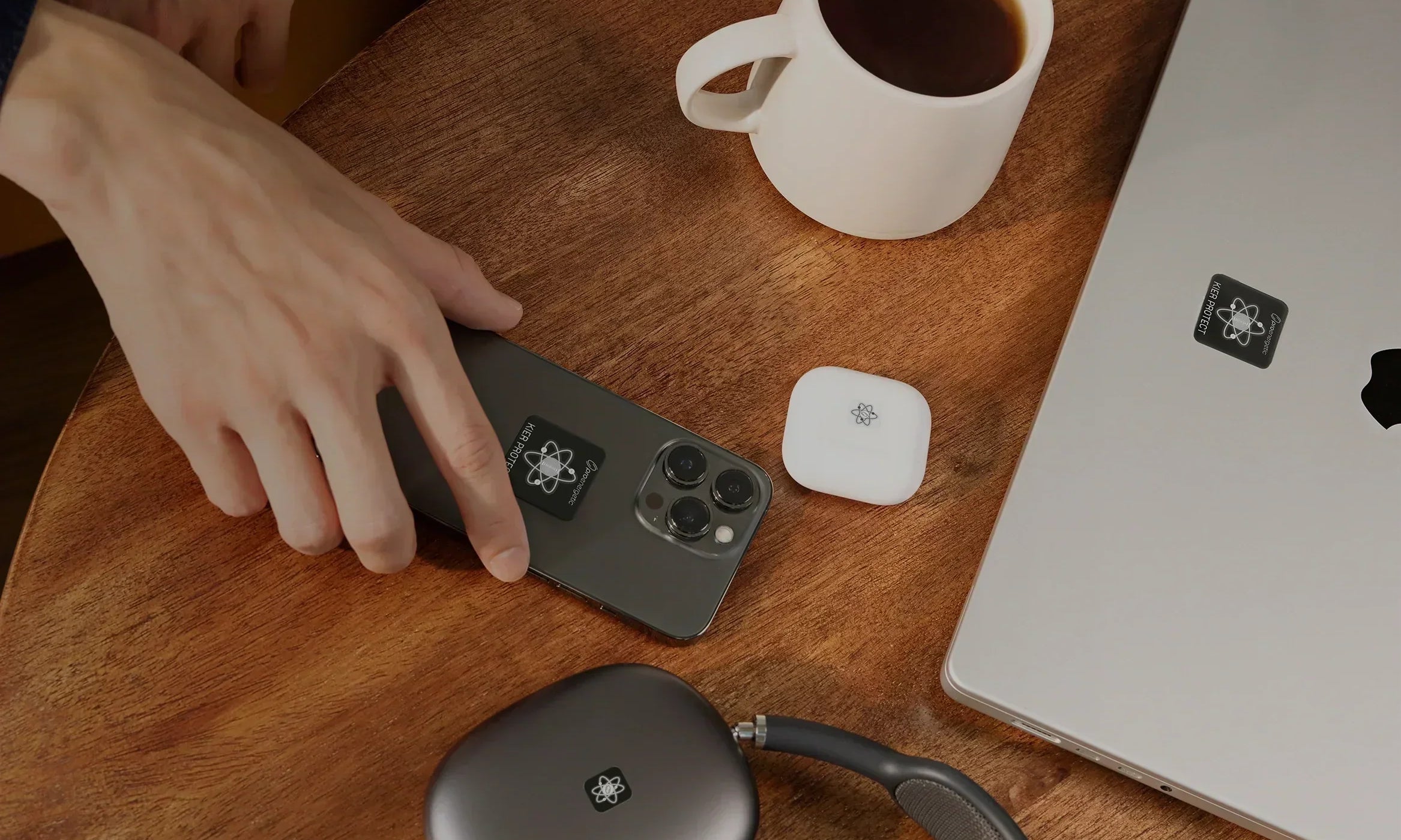 Hand auf Smartphone mit sichtbaren EMF Schutzaufklebern auf Holztisch neben Laptop und Kopfhörern mit EMF Schutzaufklebern sowie Kaffeetasse