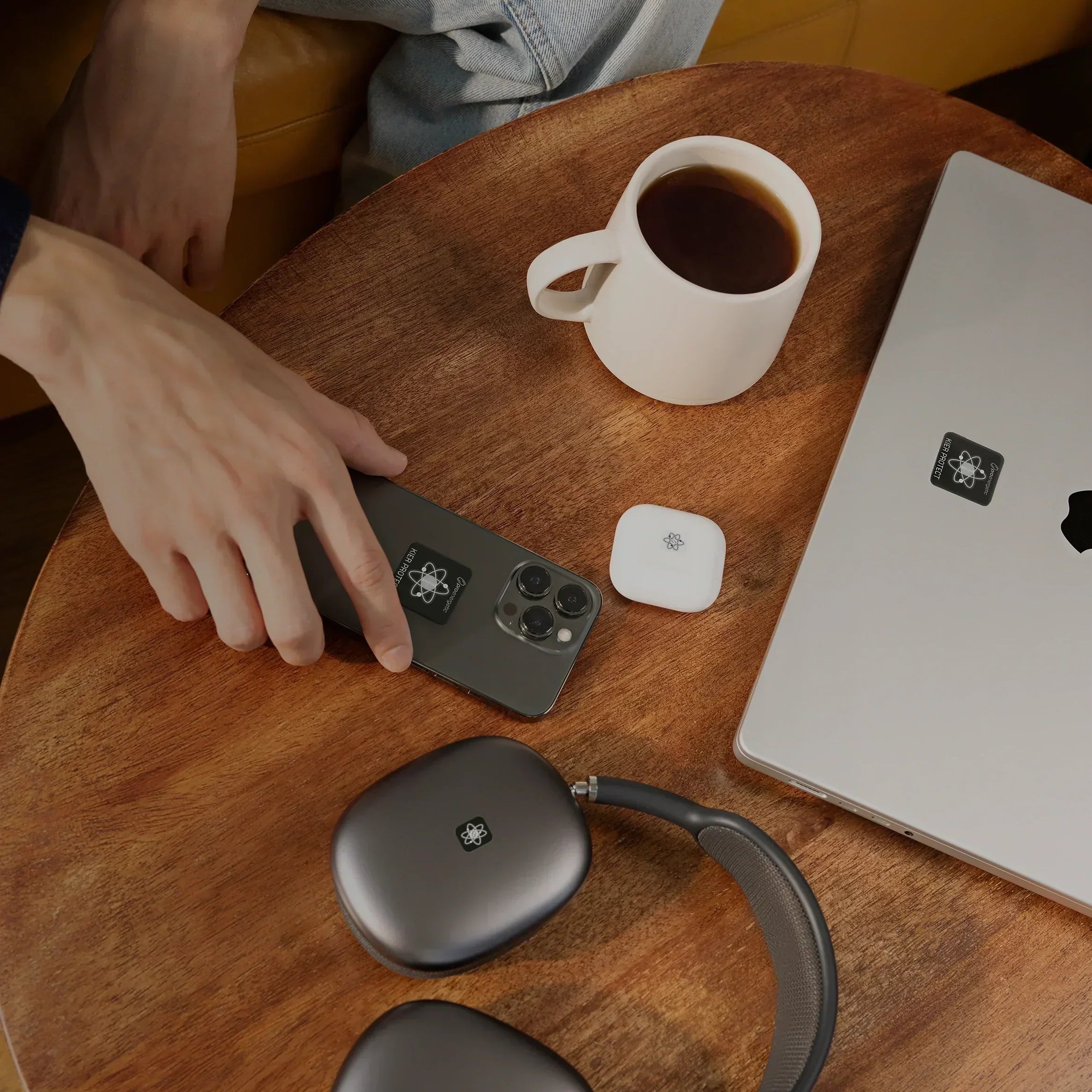 Hand auf Smartphone mit sichtbaren EMF Schutzaufklebern auf Holztisch neben Laptop und Kopfhörern mit EMF Schutzaufklebern sowie Kaffeetasse