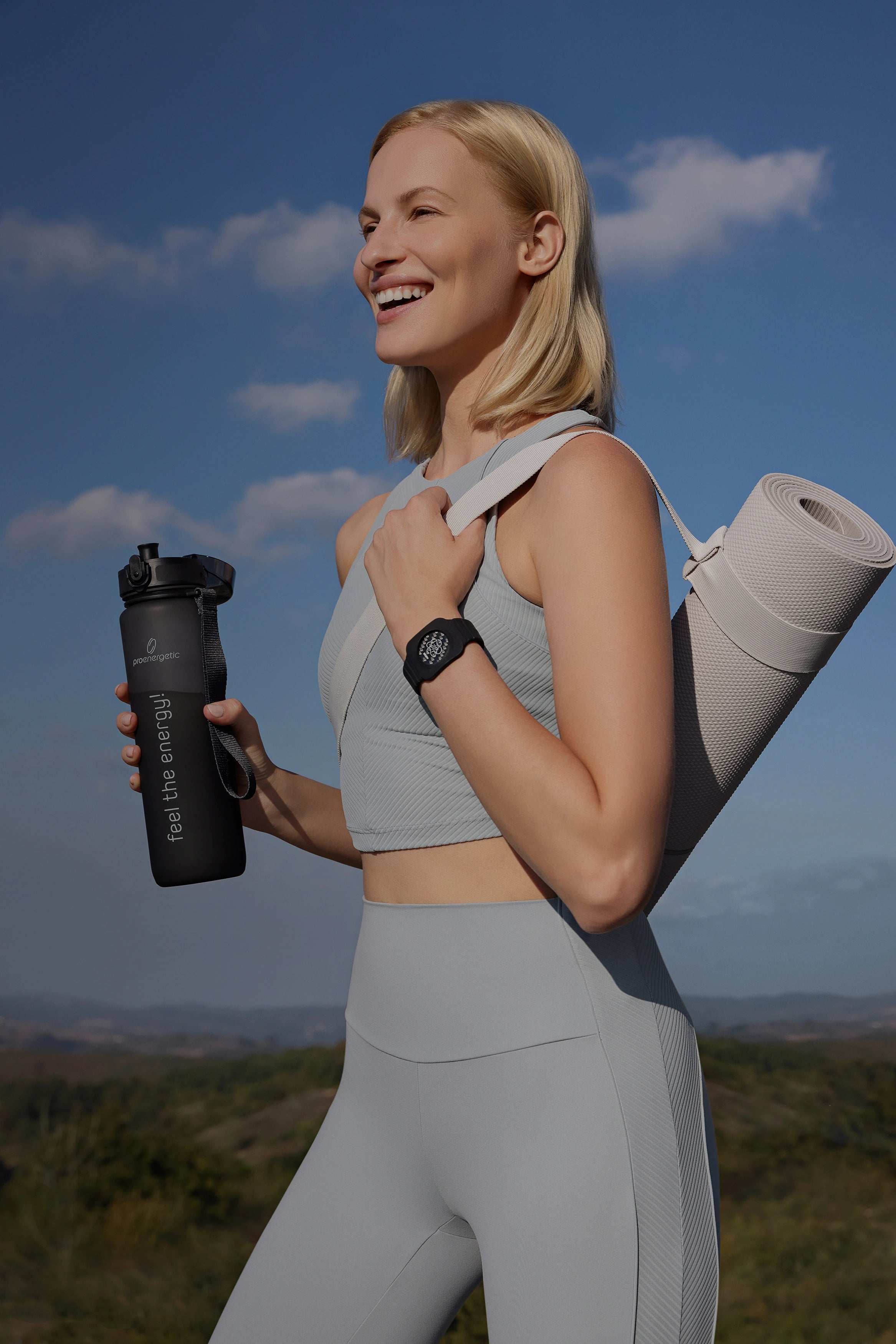 Person steht im Freien in Sportkleidung, hält die schwarze Pure Move Trinkflasche und trägt ein schwarzes Eloop Band mit Move Chip am Handgelenk vor blauem Himmel