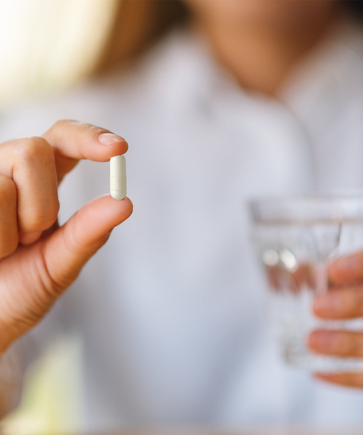 Hand hält eine einzelne helle Amino-Kapsel zwischen Daumen und Zeigefinger, im Hintergrund unscharf ein Glas Wasser.