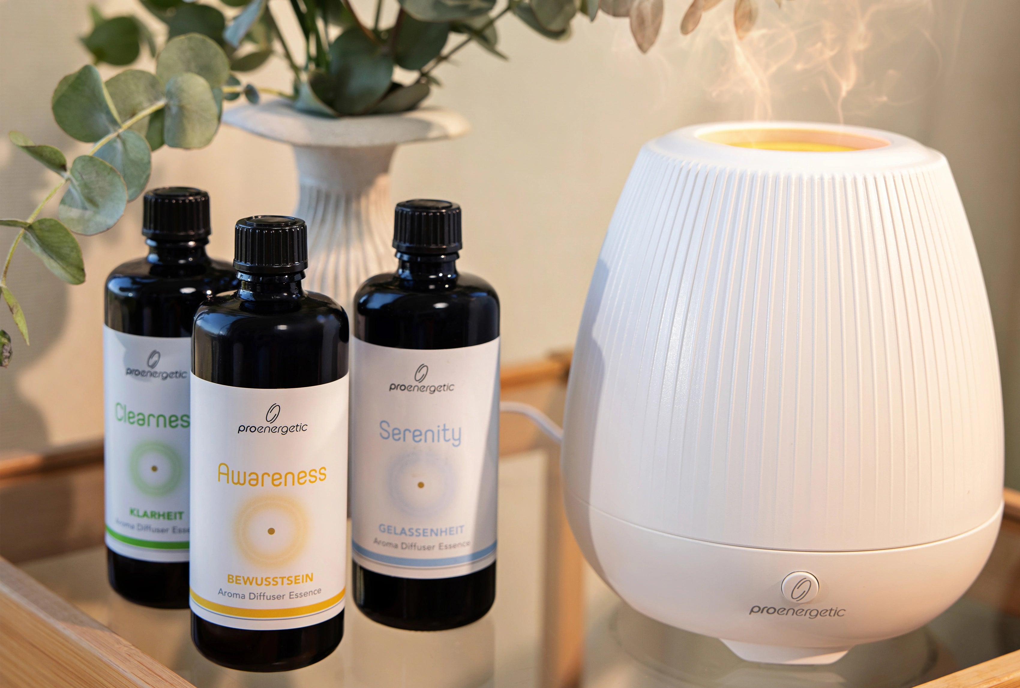Weißer ProEnergetic Aroma Diffuser auf einem Tisch, daneben drei dunkle Glasflaschen mit ProEnergetic Duftessenzen und eine Vase mit grünen Zweigen im Innenraum