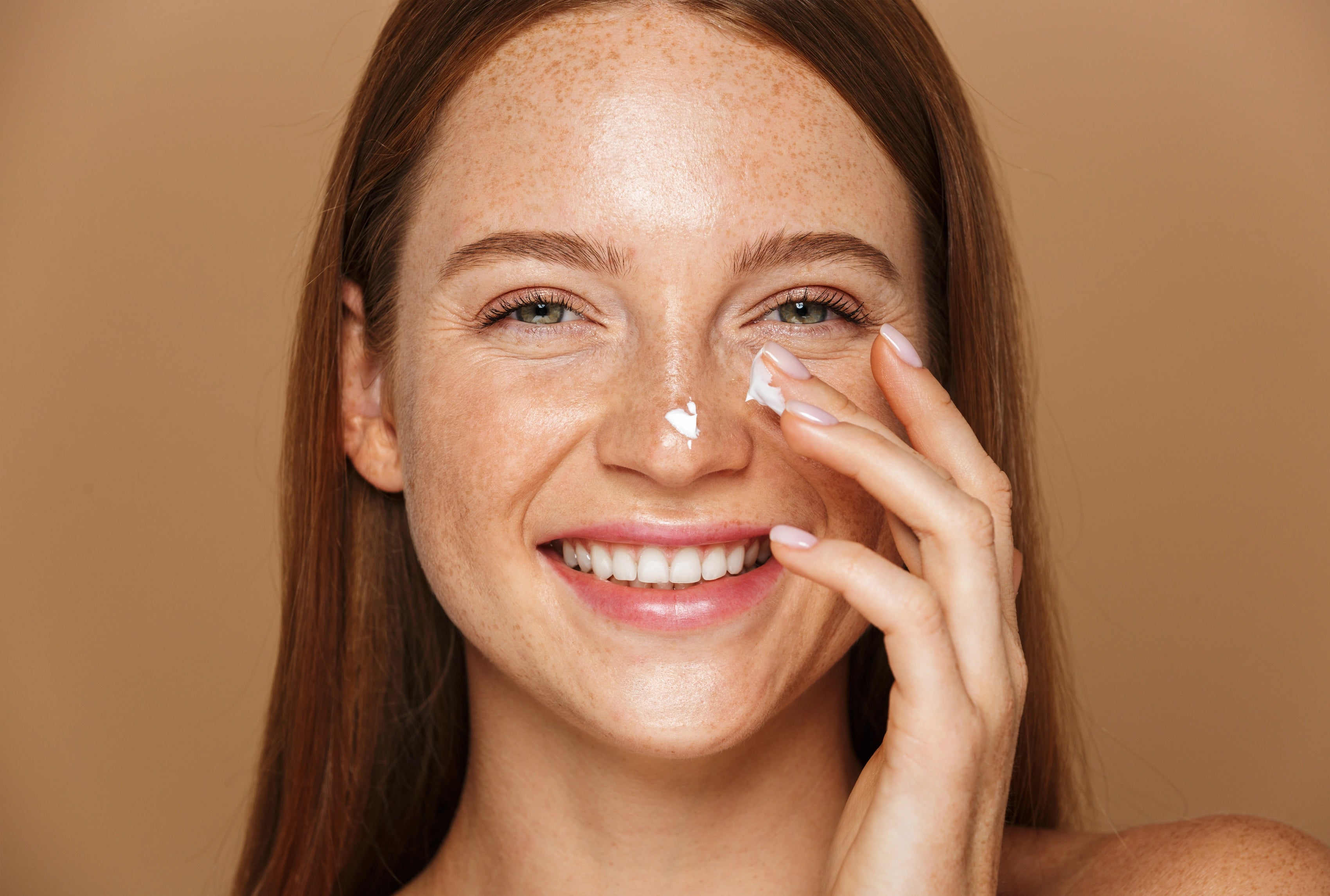 Lächelnde Frau mit Sommersprossen trägt den „Clean Skin“ Cleanser auf die Gesichtshaut auf, Creme punktuell auf Nase und Wange, vor warmem, neutralem Hintergrund.