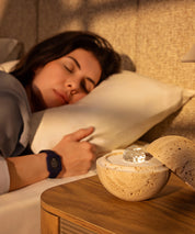 Eine Frau schläft friedlich, während sie das Eloop-Band mit sleep-Chip trägt; auf dem Nachttisch steht der Dormea aus natürlichem Sandstein.