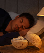 Der Dormea aus Sandstein auf einem Nachttisch, während im Hintergrund ein Mann friedlich schläft.