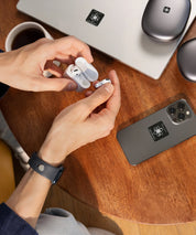 Hände nehmen kabellose In-Ear-Kopfhörer aus einem Ladecase. Auf dem Holztisch liegen Smartphone und Laptop, beide mit E-Protect-Audio-Aufklebern versehen, daneben eine Kaffeetasse.