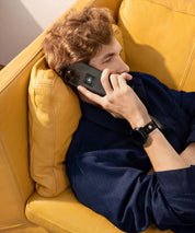 Mann liegt im Innenraum auf einem gelben Sofa, telefoniert mit einem Smartphone mit sichtbaren KIER Protect Mobile Sticker und trägt eine schwarze Smartwatch mit EMF-Protection Pin am Handgelenk