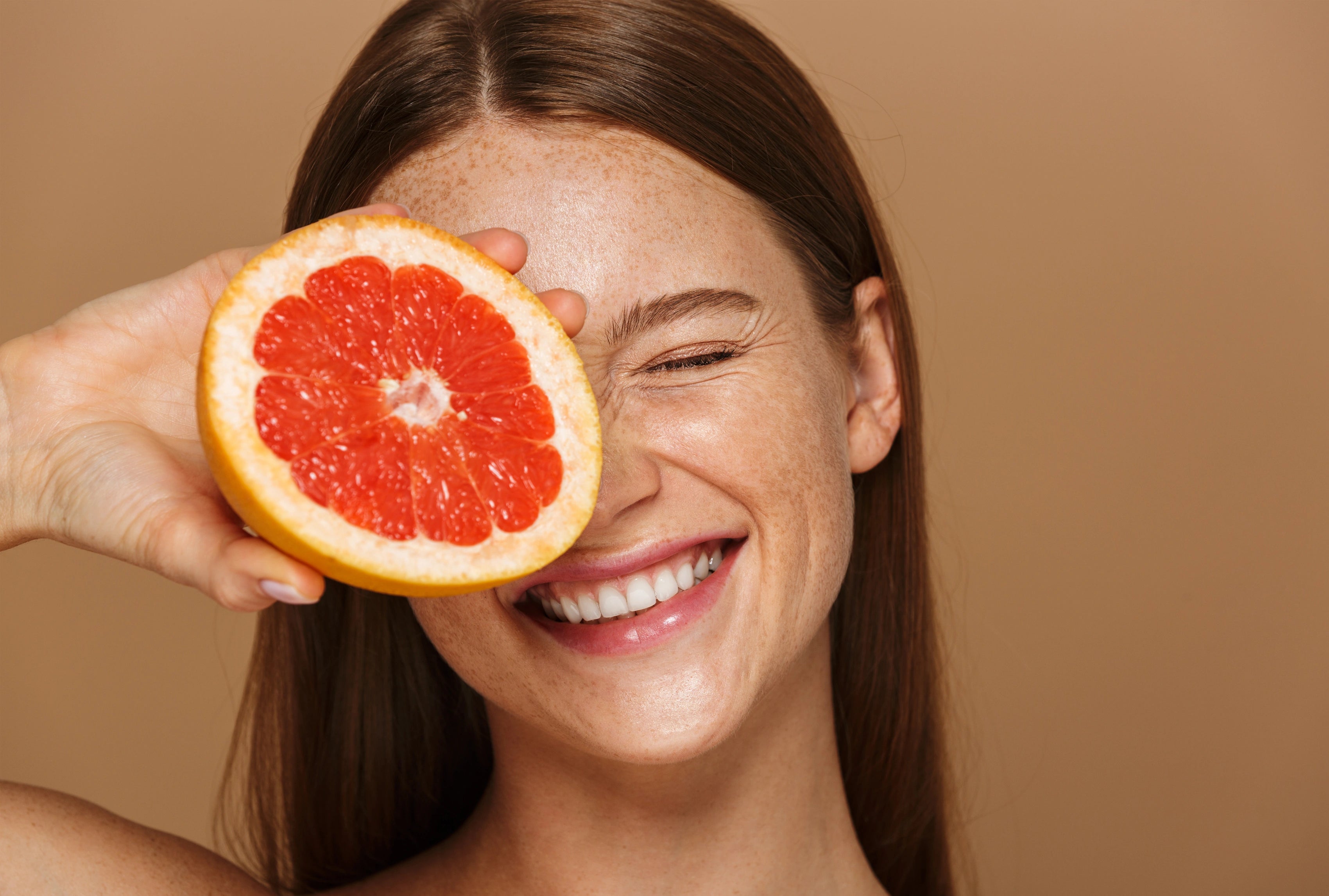 Lächelnde Frau mit Sommersprossen hält eine halbe Grapefruit vor ein Auge; frisches, natürliches Beauty-Motiv vor warmem, einfarbigem Hintergrund.
