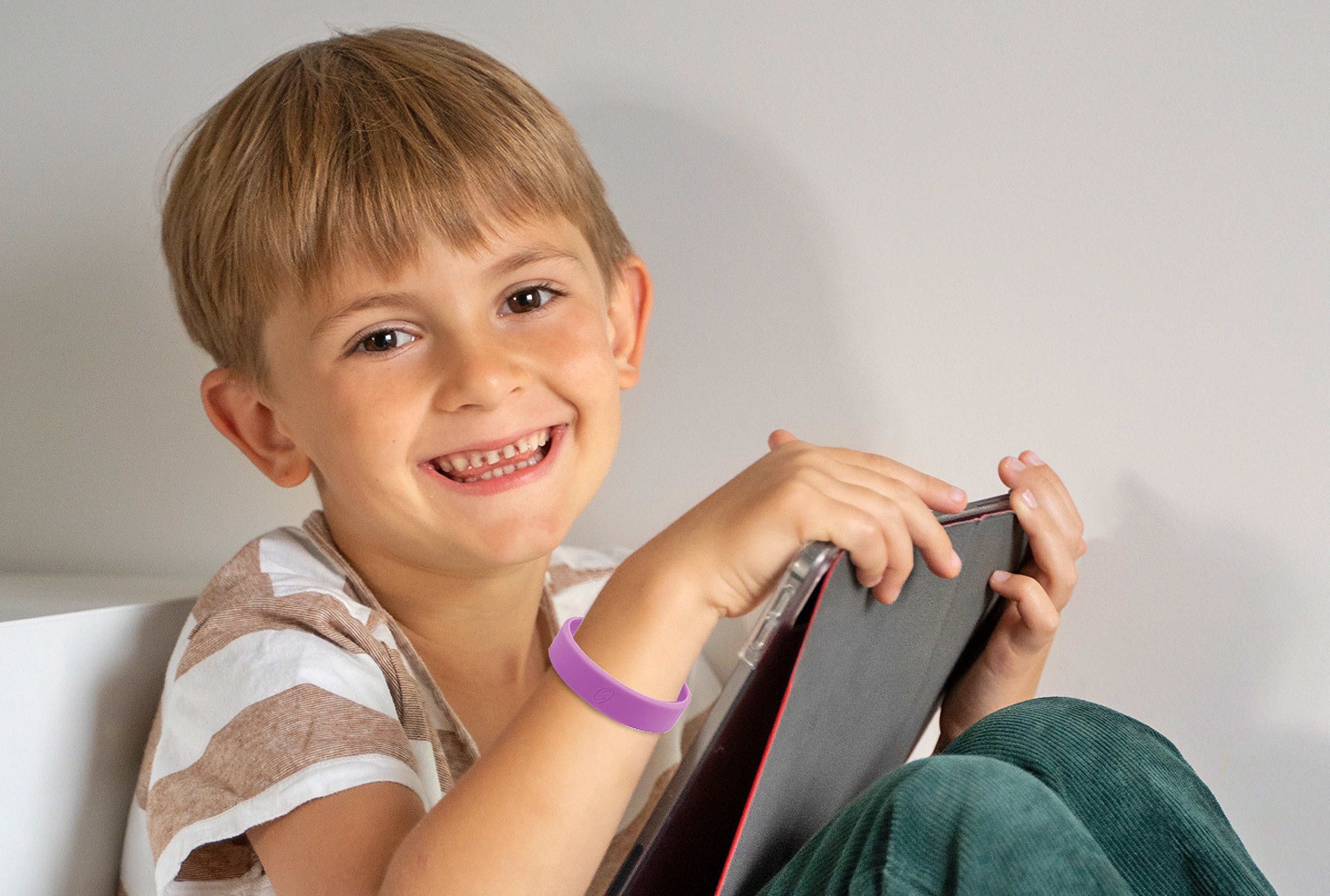 Junge sitzt im Innenraum und hält ein Tablet, am Handgelenk trägt er ein lila ProEnergetic Kids Band aus Silikon