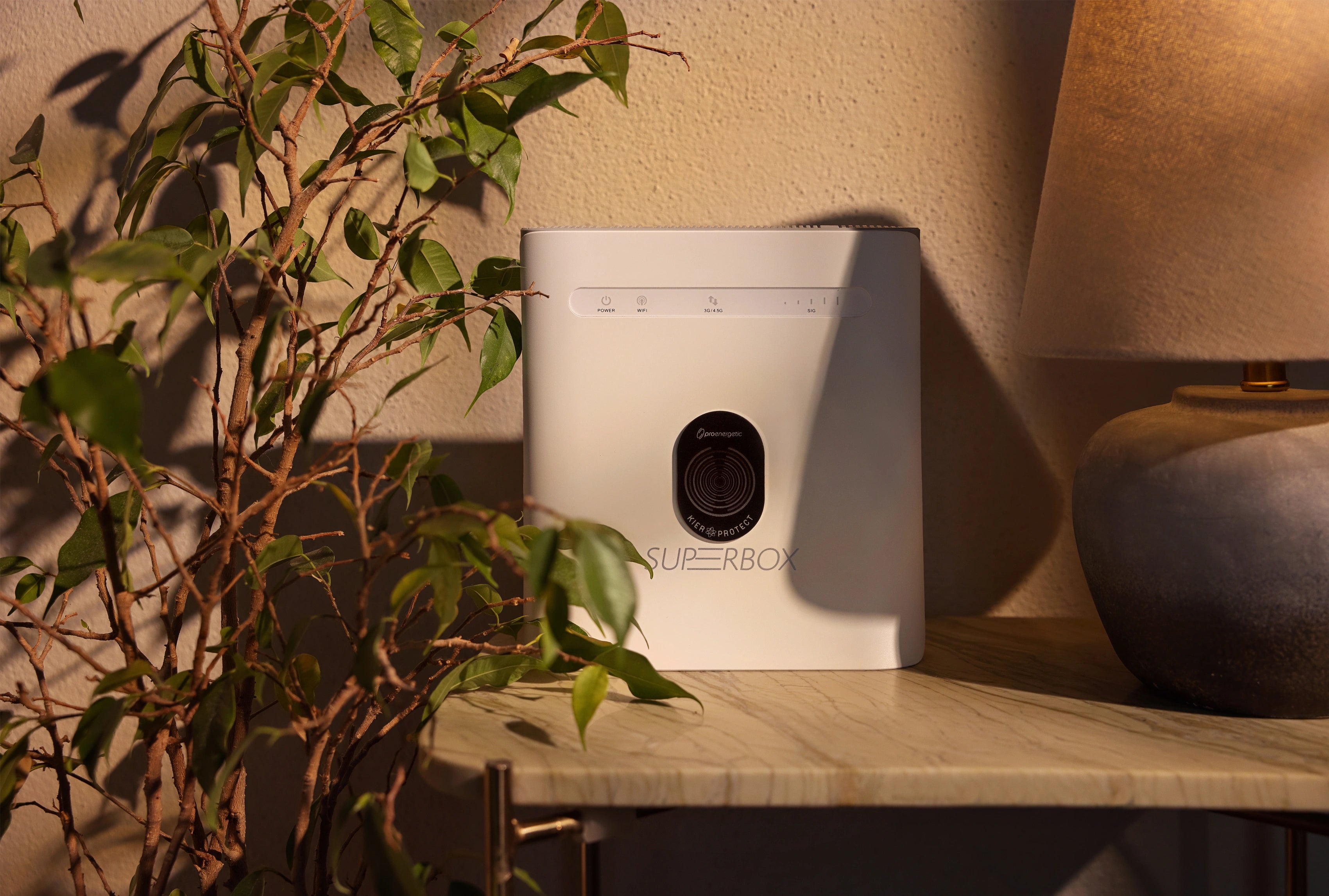 Weißes Superbox-Gerät auf einem Beistelltisch im Innenraum, schwarzes KIER Protect Home Element mittig auf der Vorderseite angebracht, Pflanze und Tischlampe sichtbar