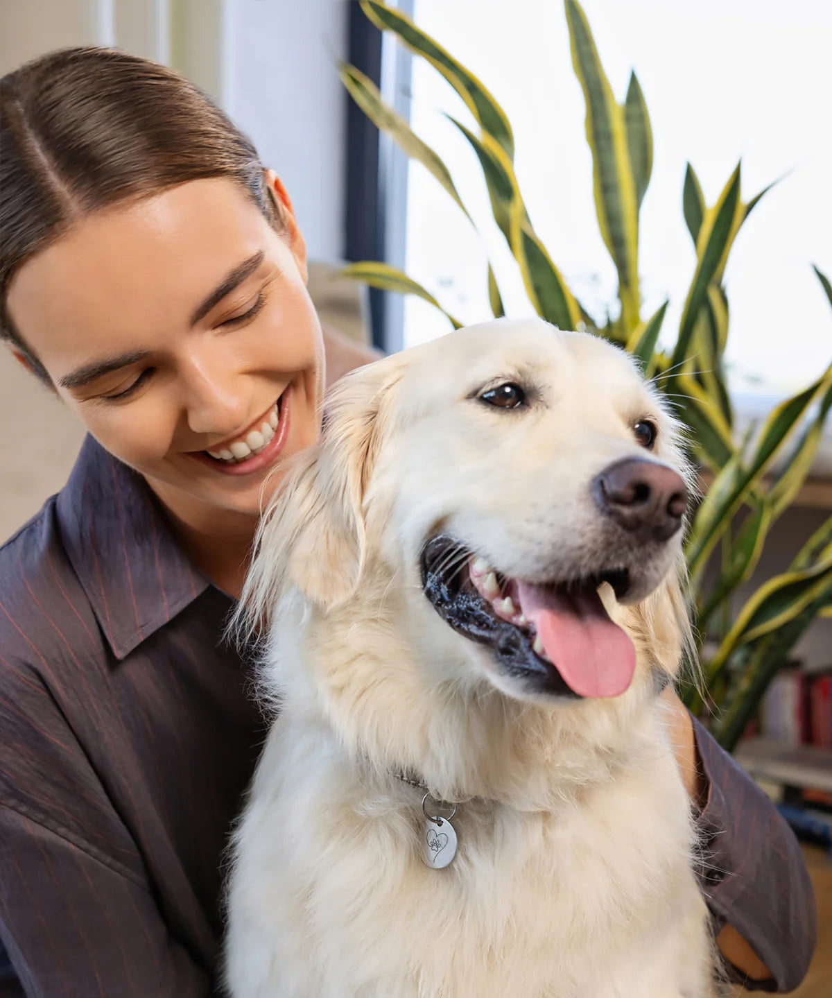 Lächelnde Frau umarmt einen hellen Golden Retriever mit silbernem Herz-Pfoten-Anhänger, dem Pet Tag Anhänger von ProEnergetic, am Halsband in wohnlicher Umgebung.