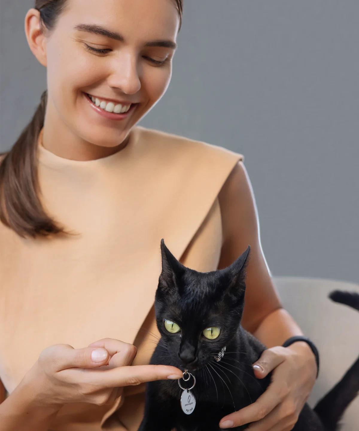Lächelnde Frau hält eine schwarze Katze auf dem Schoß und zeigt auf den silbernen Pet-Tag-Anhänger von ProEnergetic am Halsband.