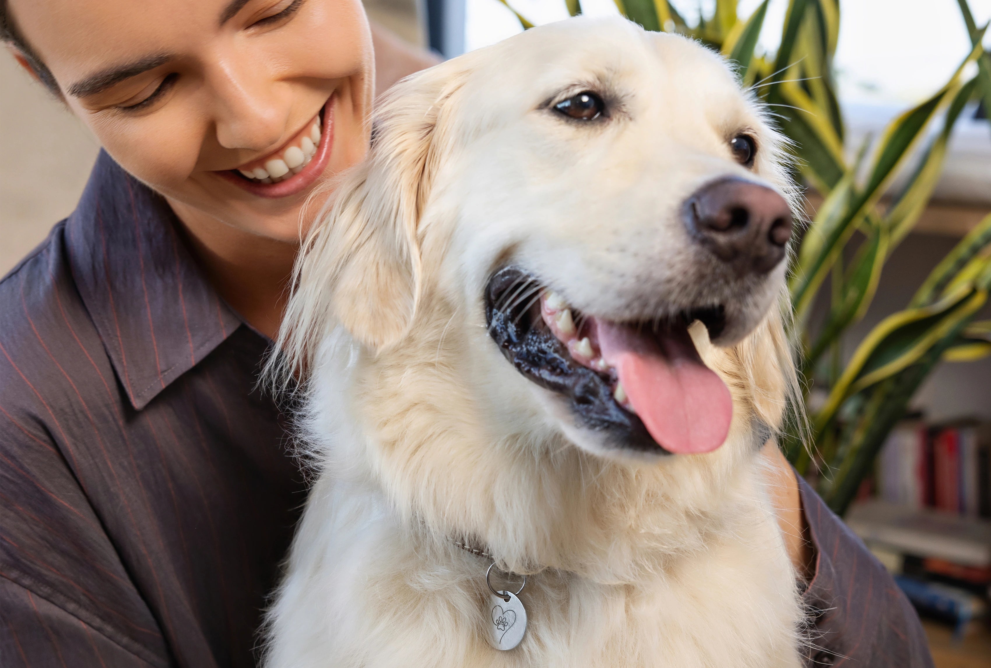 Lächelnde Frau umarmt einen hellen Golden Retriever mit silbernem Herz-Pfoten-Anhänger, dem Pet Tag Anhänger von ProEnergetic, am Halsband in wohnlicher Umgebung.