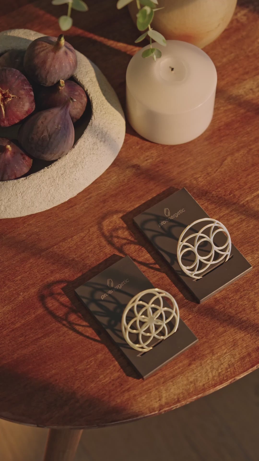 Ein Flower of Love Goodie und ein Seed of Happiness Goodie als goldene Ornamente auf schwarzen Sockeln, arrangiert auf einem Holztisch neben Kerze und Obstschale