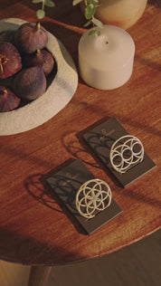 Ein Flower of Love Goodie und ein Seed of Happiness Goodie als goldene Ornamente auf schwarzen Sockeln, arrangiert auf einem Holztisch neben Kerze und Obstschale