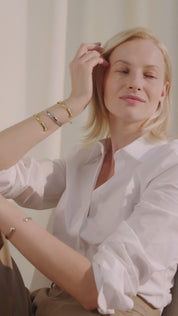 Frau sitzt in einem Innenraum auf einem Stuhl, lächelt und trägt die Lunara Cuff Armreifen in Gold und Silber mehrfach am Unterarm und Handgelenk