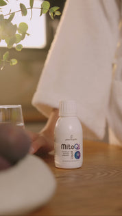 MitoQi Liquid von proenergetic: Person im hellen Bademantel gießt das braune, flüssige Supplement aus einer weißen MitoQi-Flasche in einen Löffel. Nimmt es zu sich. Natürliche, warme Lichtsituation mit Pflanzendetail im Hintergrund. Video.