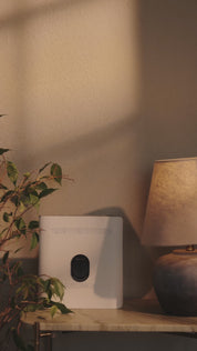 Weißes Superbox-Gerät auf einem Beistelltisch im Innenraum, schwarzes KIER Protect Home Element mittig auf der Vorderseite angebracht, Pflanze teilweise sichtbar