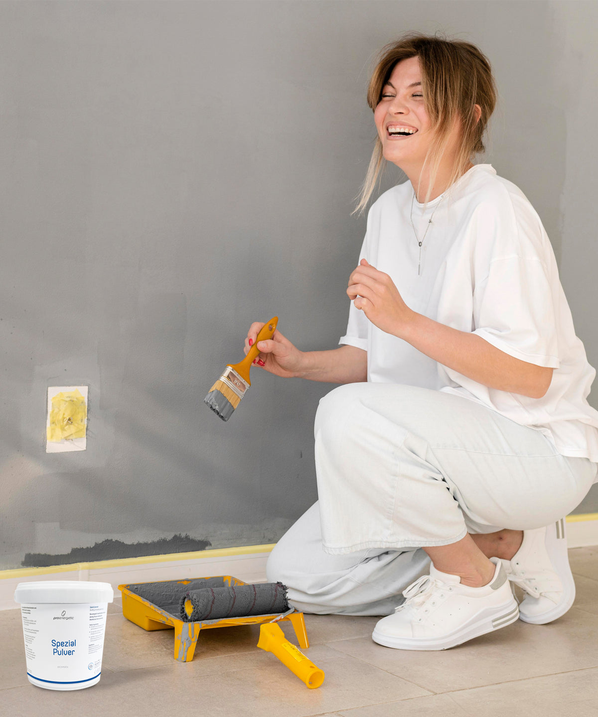 Frau kniet vor grauer Wand mit Pinsel und Farbroller neben Eimer Spezial Pulver Wohnen 800g von ProEnergetic.